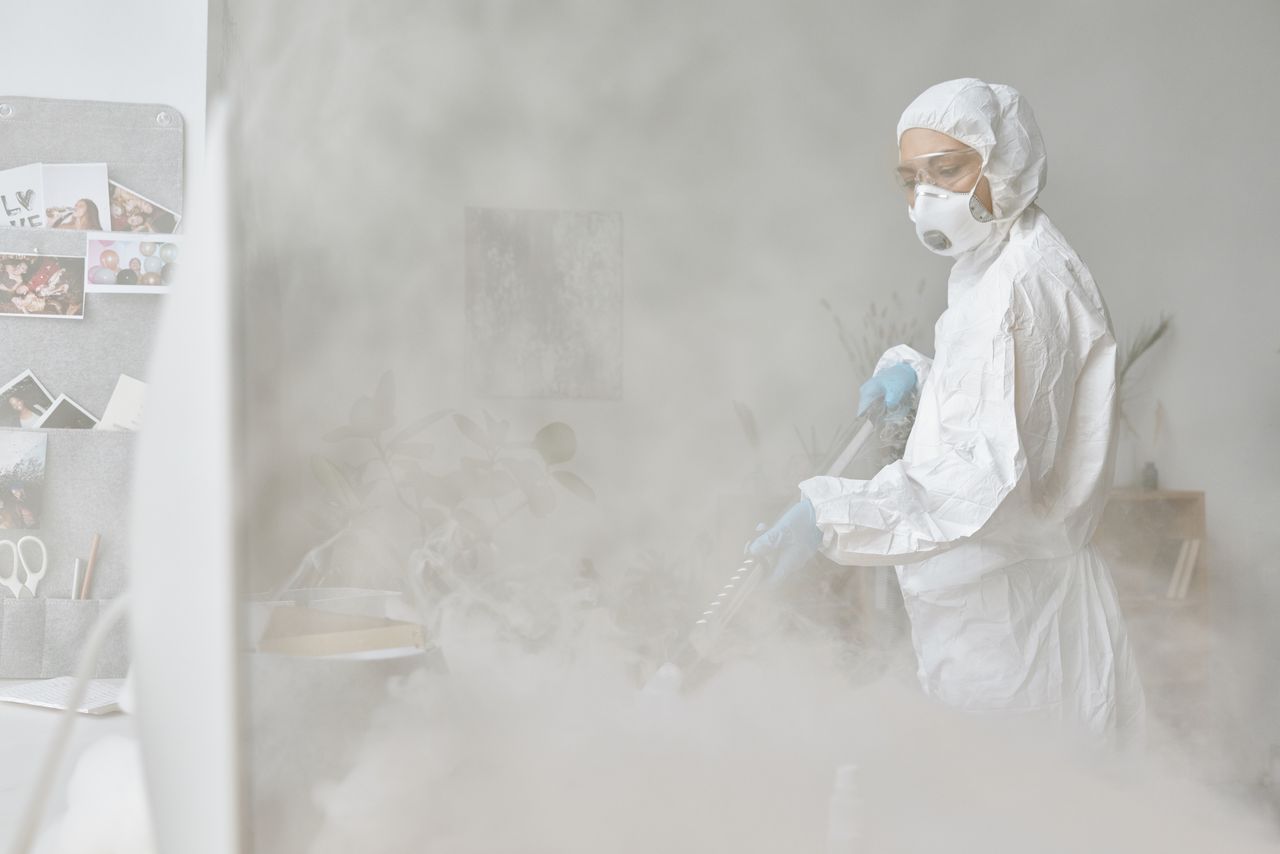 A woman in a protective suit disinfects an indoor space with fogging equipment.